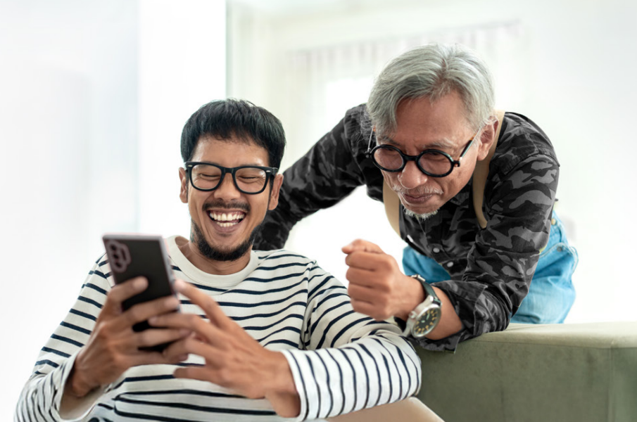 younger man laughing while showing phone to older man over his shoulder.