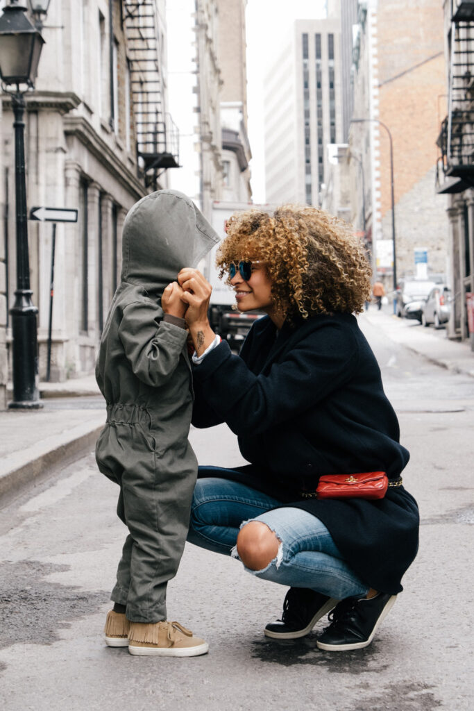 Woman with curly hair and sunglasses holding child's hands and smiling in street